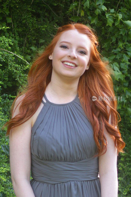 照片中，14 / 15岁的红发少女，皮肤苍白，脸上有雀斑，站在户外的花园中，穿着灰色的舞会礼服，准备迎接舞会的约会对象